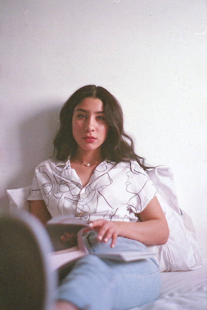 Nadya Inzunza Film Photo. A serene-looking individual with dark hair wearing a white shirt with abstract designs reclines against a white wall holding a magazine or book.